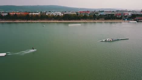 Drone-shot-of-people-wake-boarding-on-the-lake-Zlate-Piesky,-Bratislava,-Slovakia,-with-the-city-in-the-background