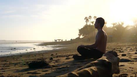 A-handsome-man-meditating-and-watching-surfers-at-sunset-on-a-picturesque-California-beach-with-palm-trees-and-people-in-silhouette
