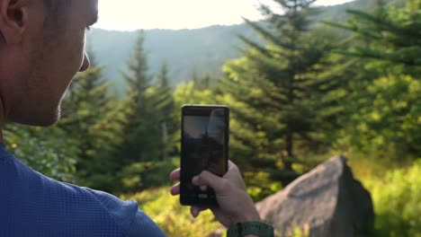 Along-a-hiking-trail,-a-young-man-pauses-to-take-a-nature-picture-with-the-multiple-cameras-on-his-smartphone