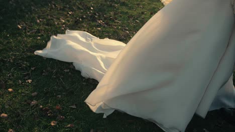 Female-white-wedding-dress-moving-in-the-wind,-on-grass-with-some-fallen-leaves,-during-sunset