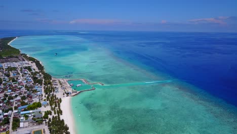 Vista-Aérea-A-Gran-Altura-De-Una-Isla-Cubierta-De-Palmeras-En-Las-Maldivas-Mientras-El-Color-Del-Mar-Va-De-Turquesa-A-Azul-Oscuro