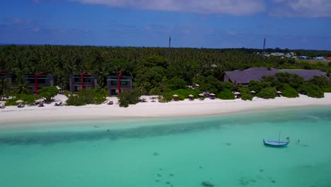 Aerial-pan-left-over-Maldives-beachfront-eco-hotel,-beautiful-crystal-clear-ocean-with-traditional-Dhoni-boat-in-bay-4k