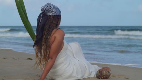 Young-female-in-a-white-beach-dress-sits-in-the-sand-while-watching-the-crashing-waves-of-the-ocean