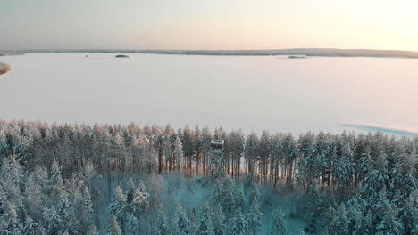 Aerial,-drone-shot,-towards-a-observation-tower,-between-snowy-pine-tree-forest,-the-snowy-lake-Pyhaselka,-of-Saimaa,-in-the-background,-on-a-sunny,-winter-evening,-in-Vuoniemi,-North-Karelia,-Finland