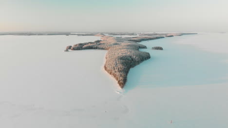Aerial,-tracking,-drone-shot,-of-Vuoniemi-cape,-in-the-middle-of-snowy-lake-Saimaa,-full-of-pine-tree-forest,-at-sunset,-on-a-sunny,-winter-evening-dusk,-in-North-Karelia,-Finland