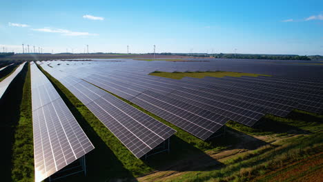 Aerial-trucking-shot-of-large-photovoltaic-solar-farm-on-field-during-blue-sky-and-sunlight---Wind-turbine-farm-in-background