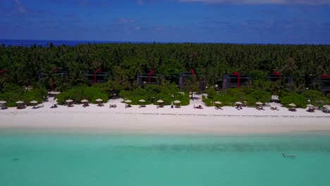 Aerial-pan,-people-swim-in-turquoise-Maldives-beach-resort-with-seaside-bungalows
