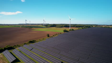 Vuelo-De-Drones-Sobre-Una-Enorme-Granja-Solar-Fotovoltaica-En-El-Campo-Con-Turbinas-Eólicas-Giratorias-Durante-El-Cielo-Azul