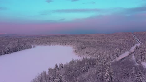 Luftaufnahme,-Die-Rückwärts-über-Einem-Verschneiten-Wald-Und-Einem-See-Mit-Rosa-Sonnenaufgang-Fliegt