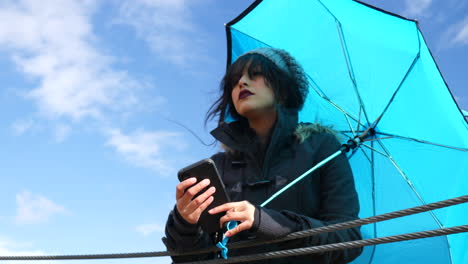 Pretty-young-woman-checking-the-weather-or-texting-on-smartphone-with-umbrella-under-blue-skies-as-rain-storm-clouds-approach