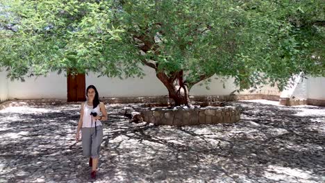 A-girl-walking-under-a-big-tree-on-a-historical-courtyard,-holding-her-camera