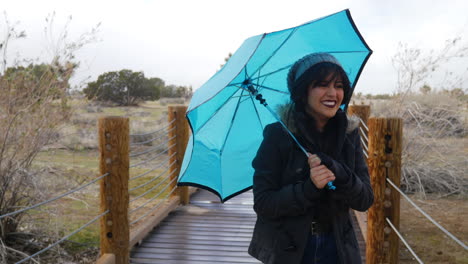 A-pretty-woman-walking-in-rain-and-windy-bad-weather-during-a-storm-with-a-blue-umbrella-as-she-smiles-and-laughs
