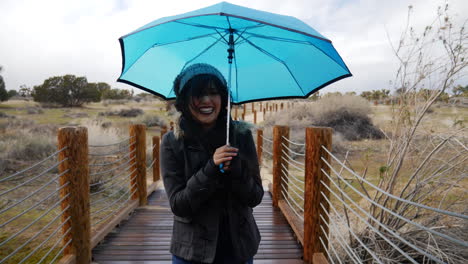 Eine-Schöne-Junge-Frau,-Die-Im-Regen-Lächelt-Und-Lacht,-Mit-Einem-Blauen-Regenschirm-Im-Sturm-Und-Bei-Schlechtem-Wetter-In-Zeitlupe