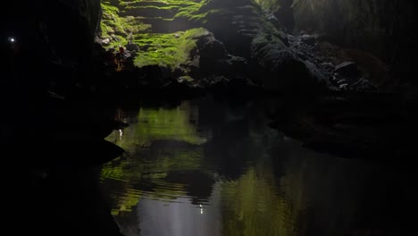 Pan-upward-from-the-reflection-on-the-water-to-the-doline-with-sunbeam-inside-SON-DOONG-cave,-people-silhouettes-moving