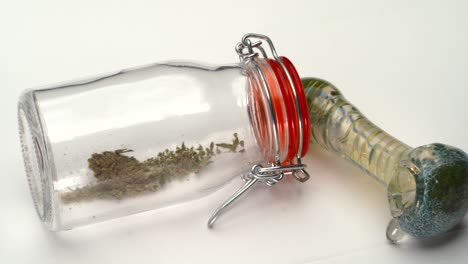 Pan-across-a-clear-bottle-with-marijuana-grounds-inside-and-glass-smoking-pipe