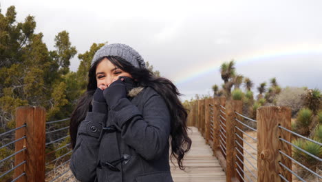 Attractive-young-woman-smiling-in-a-cold-winter-rain-storm-with-a-colorful-rainbow-in-the-cloudy-sky-SLOW-MOTION