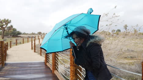 A-beautiful-woman-in-bad-weather-with-a-blue-umbrella-during-the-strong-winds-of-a-rain-storm-in-the-desert-SLOW-MOTION