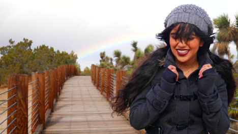A-happy,-beautiful-young-woman-walking-through-the-desert-with-a-rainbow-in-the-cloudy-sky-during-a-rain-storm-SLOW-MOTION