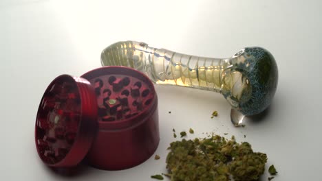 Closeup-view-of-a-glass-pipe-on-a-white-table-as-the-camera-pulls-back-to-reveal-an-open-red-grinder-and-marijuana-buds