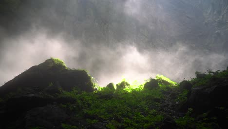 Statischer-Nebelstrahl,-Der-Mit-Dem-Sonnenstrahl-Schwebt,-Der-Auf-Felsen-Scheint,-Die-Mit-Grünen-Pflanzen-In-Einer-Höhle-Bedeckt-Sind