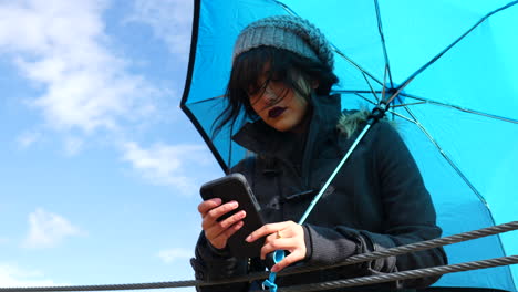 Attraktive-Junge-Frau,-Die-Auf-Dem-Smartphone-SMS-Schreibt-Und-Wetterschirm-Unter-Blauem-Himmel-Hält,-Während-Regensturmwolken-Vorbeiziehen