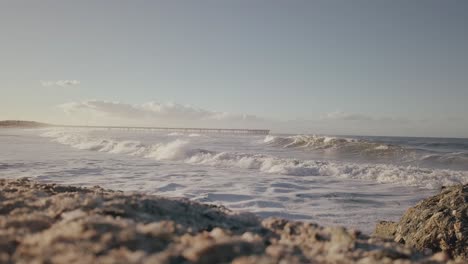 Close-up-slow-motion-sandy-beach,-rolling-waves-with-view-of-Venice-beach-pier-across-the-water-in-distance-on-sunny-day