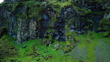 Aerial-landscape-view-of-a-dark-mountain-cliff,-covered-in-green-moss,-in-Iceland,-with-birds-flying-by