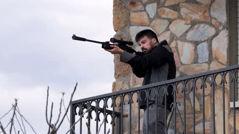Young-man-with-beard-dressed-in-black-looks-through-the-gun-sight-of-a-hunting-rifle-in-a-balcony