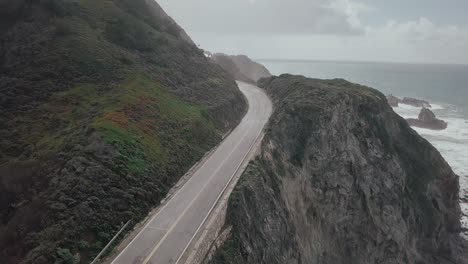 Aerial-drone-view-across-South-Pacific-Highway-following-coastal-route-with-views-of-the-ocean-waves-alongside-rocky-terrain