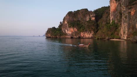 aerial-drone-pull-away-shot-near-Railay-Beach-in-Krabi-Thailand-during-sunrise-as-a-Thai-longtail-boat-motors-through-the-Andaman-Sea-with-a-tourist-onboard-and-large-limestone-mountains-behind
