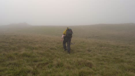Mann-In-Gelber-Jacke,-Der-Einen-Grasbewachsenen-Hügel-Hinauf-In-Den-Nebel-Wandert