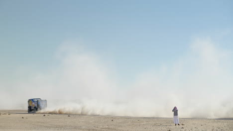 Ein-Blauer-Dakar-rallye-truck,-Der-An-Einem-Arabischen-Mann-Vorbeirast,-Der-Fotos-Mit-Staubwolken-Macht