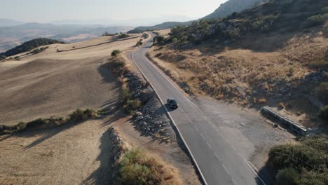 Aerial-drone-shot-over-a-black-car-while-passing-through-rural-hilly-landscape-in-El-Torcal-de-Antequera,-Sierra-in-Spain-during-evening-time
