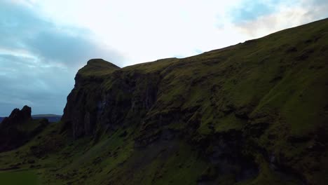 Aerial-panoramic-landscape-view-of-a-dark-mountain-cliff,-covered-in-green-moss,-in-Iceland,-at-dusk