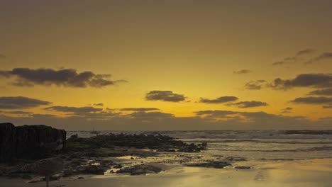 Kleine-Wellen-Umspülen-Eine-Buhne-An-Einem-Strand,-Während-Der-Himmel-Vom-Sonnenuntergang-Orange-Gefärbt-Wird