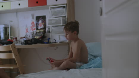 Seven-years-old-boy-sitting-on-bed,-in-his-bedroom-and-using-a-digital-table,-in-daylight