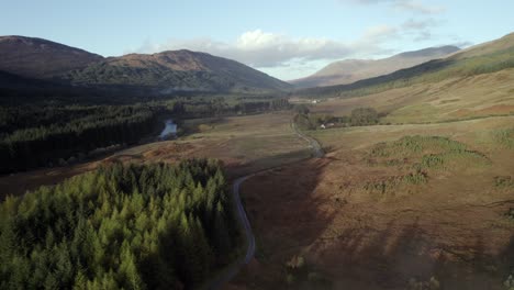 Aerial-drone-footage-of-a-winding-single-track-road-stretching-out-towards-mountains,-surrounded-by-a-forest-of-conifer-trees,-moorland-and-a-river