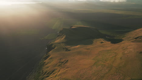 Slow-aerial-pullout-above-golden-cliff-near-black-sand-beach-at-sunset,-Iceland
