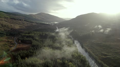 Aerial-drone-footage-flying-high-and-slowly-above-a-river,-road-and-forest-of-conifer-trees-while-low-hanging-cloud-hugs-the-treetops-and-the-sun-sets-behind-mountains