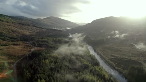 Aerial-drone-footage-flying-high-above-a-river,-road-and-forest-of-conifer-trees-while-low-hanging-cloud-hugs-the-treetops-and-the-sun-sets-behind-mountains