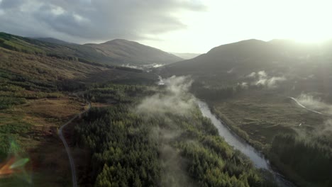 Aerial-drone-footage-flying-slowly,-high-above-a-river,-road-and-forest-of-conifer-trees-while-low-hanging-cloud-hugs-the-treetops-and-the-sun-sets-behind-mountains