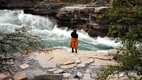 Female-Hiker-Standing-Near-Ledge-Taking-Photo-Of-Raging-Flowing-River-Water-In-Sweden