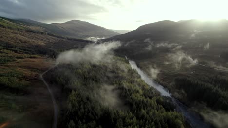 Aerial-drone-footage-high-above-the-canopy-of-a-conifer-forest-and-river-slowly-descends-as-low-hanging-cloud-hugs-the-treetops-and-the-sun-sets-behind-mountains-on-the-horizon