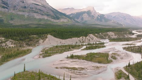 Aerial-orbit-of-river-streams-flowing-between-pine-tree-forest,-Canadian-Rockies-in-background,-Banff-National-Park,-Alberta,-Canada