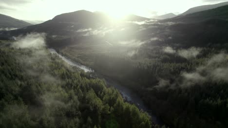 Aerial-drone-footage-slowly-rising-above-a-river-and-a-forest-of-conifer-trees-while-shafts-of-light-illuminate-low-hanging-cloud-in-the-treetops-and-the-sun-sets-behind-mountains