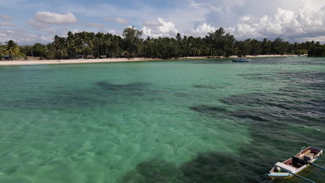 Barco-Navegando-En-El-Agua-Cristalina-Cerca-De-La-Playa-Con-Gente-Nadando-En-Un-Día-Soleado-En-La-Isla-De-Sumba,-Indonesia