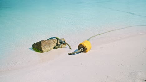 Stone-Tied-With-Thick-Rope-Used-As-Anchor-Near-Yellow-Floating-Tool-On-Sandy-Beach