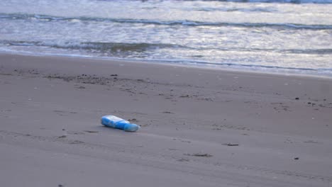 Blue-plastic-bottle-on-the-beach,-trash-and-waste-litter-on-an-empty-Baltic-sea-white-sand-beach,-environmental-pollution-problem,-calm-evening,-medium-wide-shot