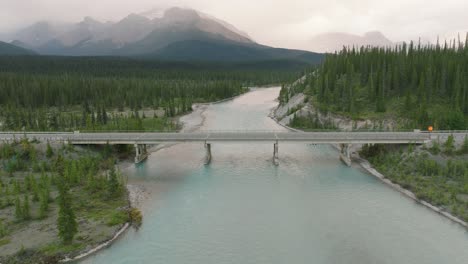 Aerial-dolly-in-of-road-bridge-over-Bow-River-between-pine-tree-forest,-Canadian-Rockies-in-back-at-Banff-National-Park,-Alberta,-Canada