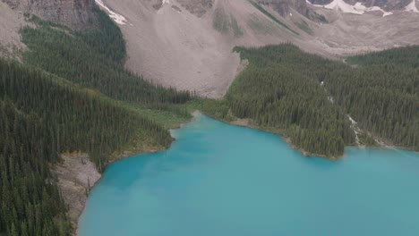 Aerial-dolly-in-lowering-on-emerald-Lake-Moraine,-pine-tree-forest-and-mountains-at-Banff-National-Park,-Alberta,-Canada